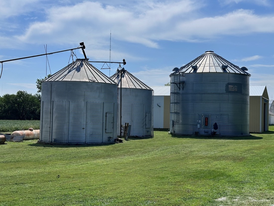 Three steel grain bins on mowed grass with a machine shed behind.
